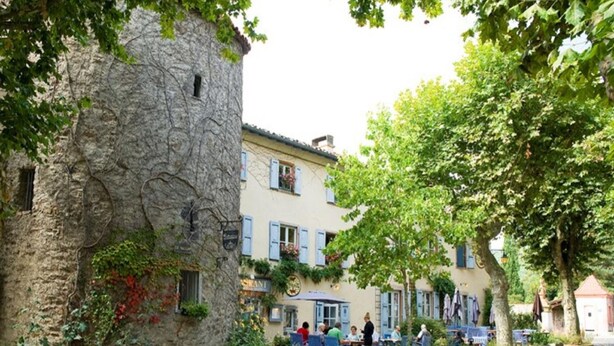 A stone building with a café patio, outdoor seating, and a tree-lined street.