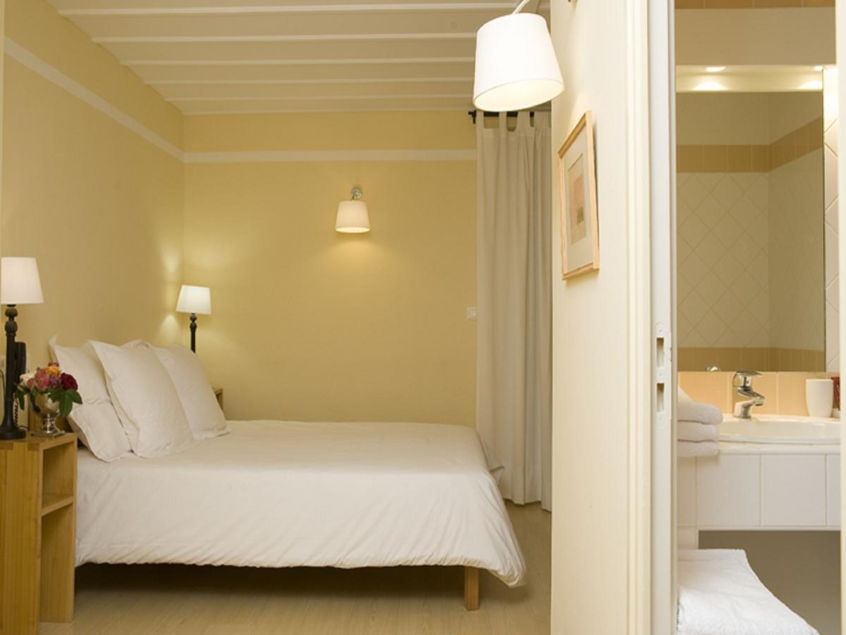 Une chambre d’hôtel avec un lit, des lampes de chevet, une table de chevet ornée d’un vase de fleurs, un tableau encadré au mur et une salle de bain visible à travers une porte ouverte.