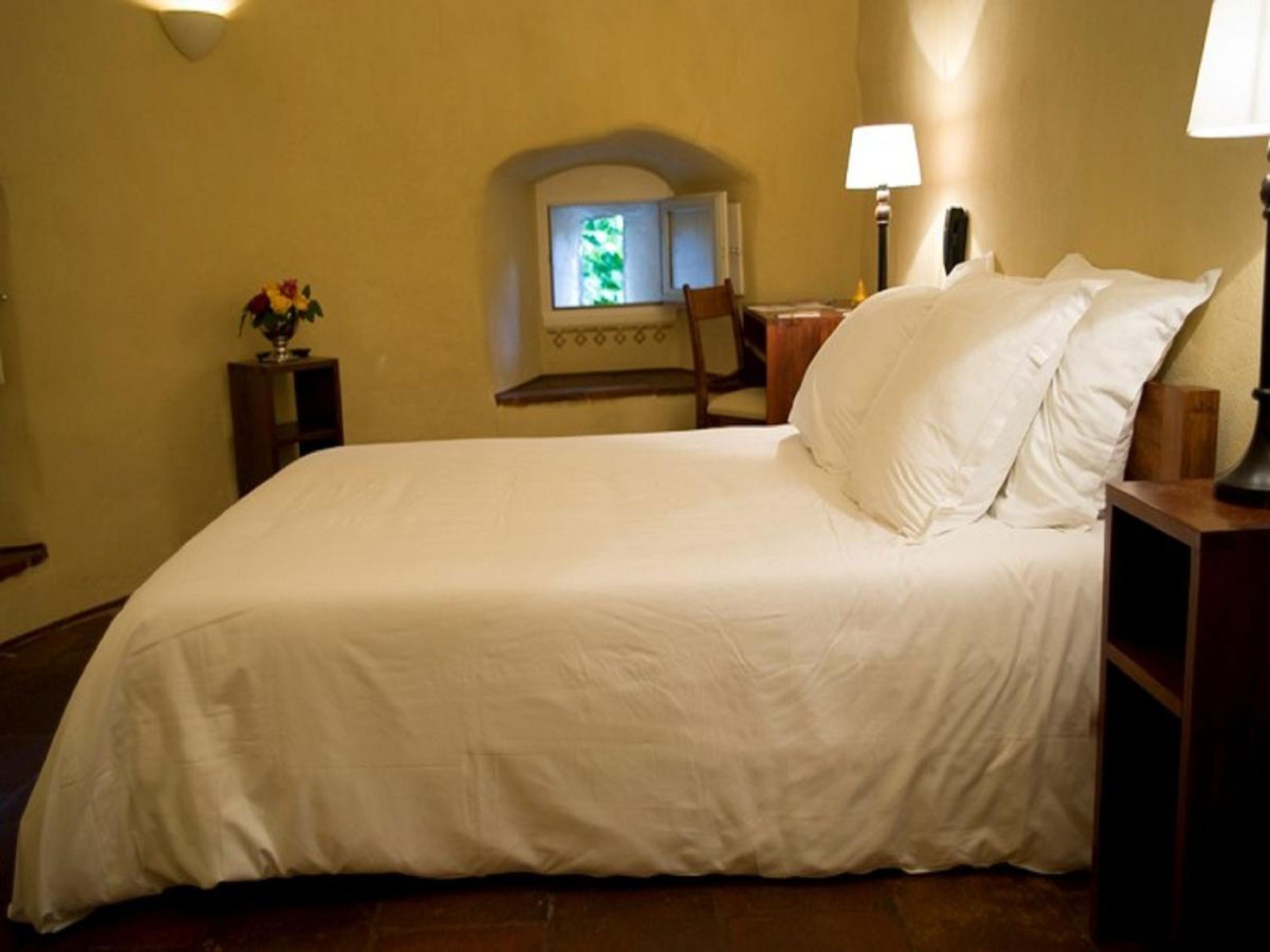 A neatly made bed in a hotel room with a small table and chair, a lamp, and a window with a view of greenery.