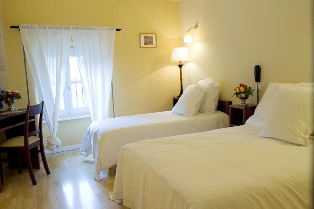 A hotel room with two beds, a desk, and a chair. There is a window with curtains, a bedside lamp, and a wall-mounted light.