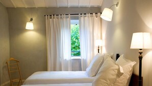 A double bed with white linens, two bedside lamps, a window with white curtains, and a wooden ladder against the wall.