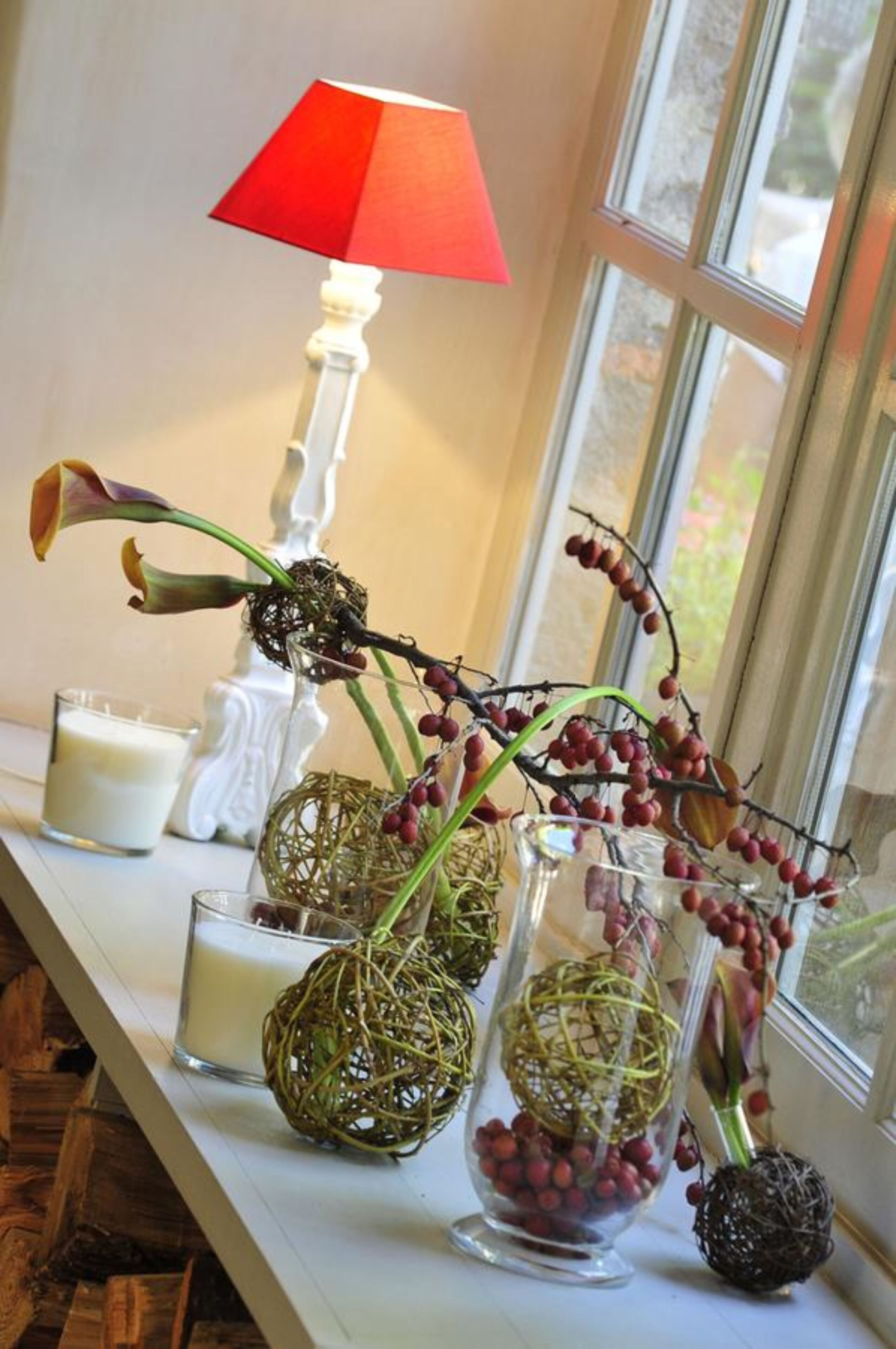 A window with a red lamp, a white vase with red berries, and a glass vase with a spherical object.