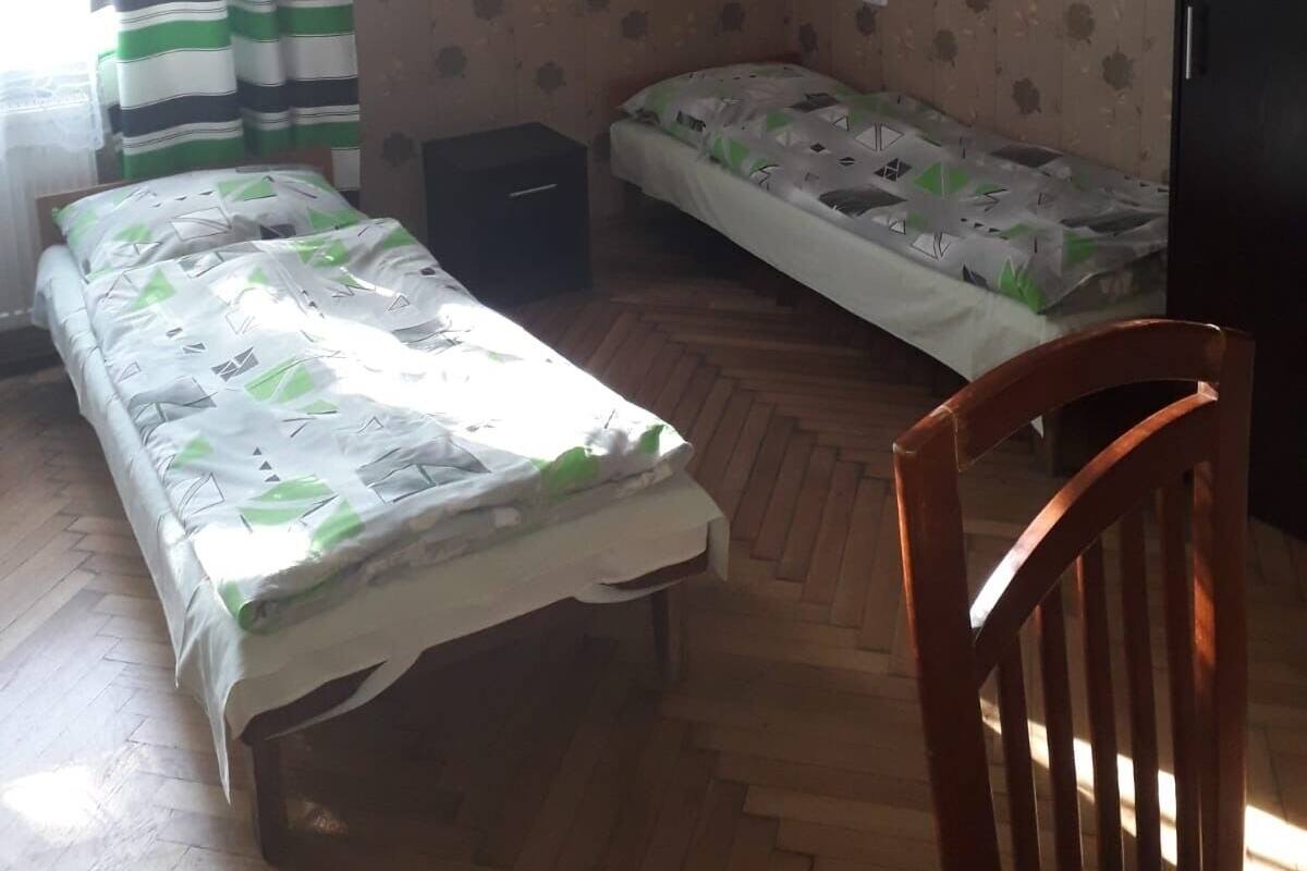 A small, sparsely furnished room with two single beds, a wooden chair, and a small dark-colored cabinet.