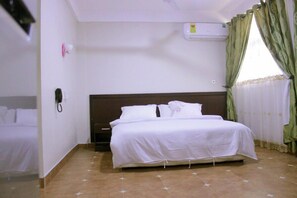 A hotel room with a single bed, a bedside table, a wall-mounted phone, and a window with green curtains.