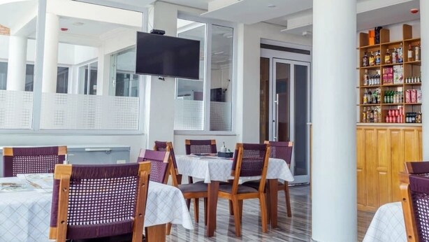 A modern dining area with tables and chairs, a flat-screen TV, and a kitchen area with shelves of products. - Royal Baron Hotel (Kumasi)