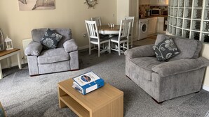 A living room with two grey sofas, a wooden coffee table, a dining table with white chairs, a kitchen area, and a large window with a view of the beach. - Harbour View Luxury Comfortable Holiday Apartment (Donaghadee)