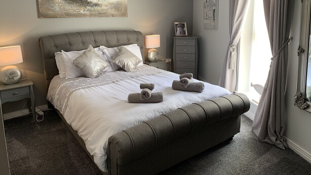 A neatly arranged bedroom with a large bed, two bedside tables, a nightstand, a dresser, and a chandelier. - Harbour View Luxury Comfortable Holiday Apartment (Donaghadee)