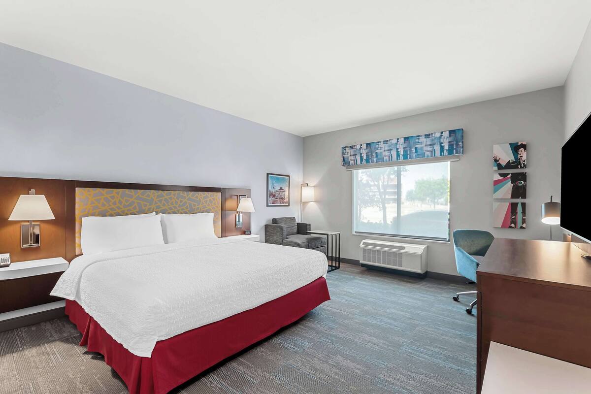 A hotel room with a large bed, a desk, a chair, a window, and a wall-mounted TV.