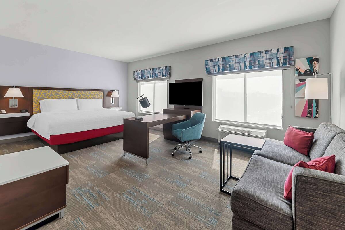 A hotel room with a bed, desk, chair, sofa, TV, and artwork on the walls.