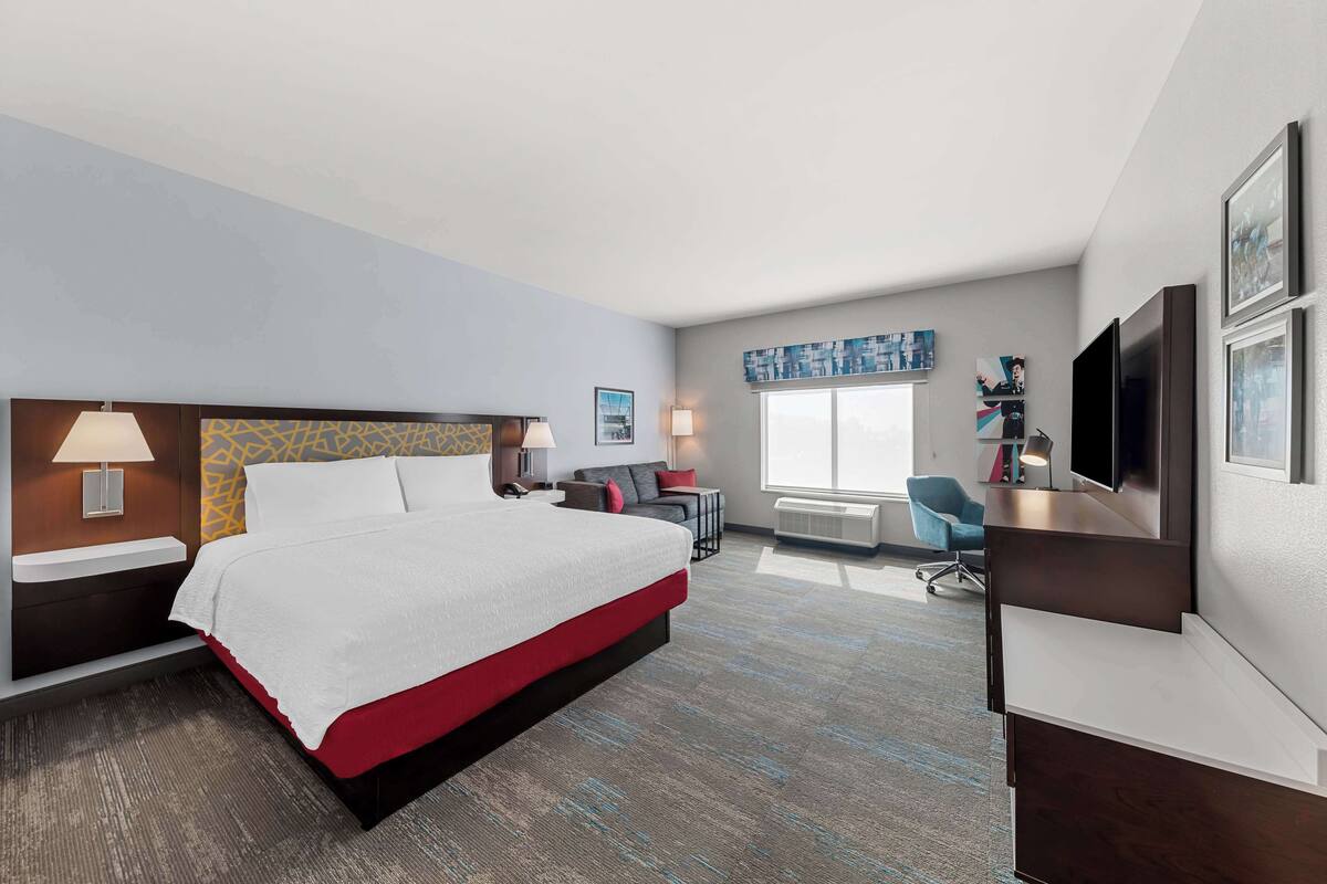 A hotel room with a large bed, bedside tables, a desk, a chair, a TV, and artwork on the walls.