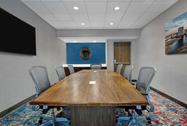 A conference room with a large wooden table, six chairs, a TV mounted on the wall, a blue accent wall, and a painting of a cityscape.