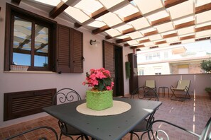 A covered outdoor patio with a table and chairs, a potted plant, and a window with shutters. - Casa Levante (Oliveri)