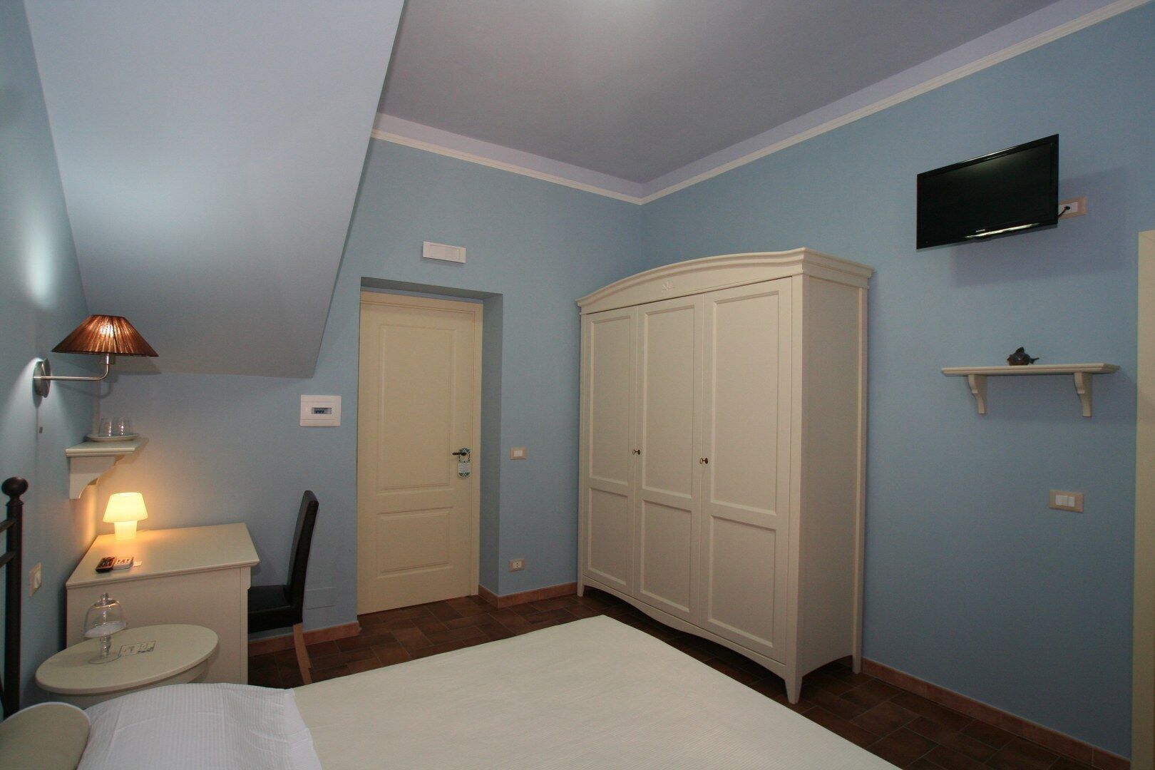 Un dormitorio con cama, mesita de noche, lámpara, armario y televisor montado.