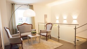 Een woonkamer met een bank, twee fauteuils, een bijzettafeltje met een plant, een vloerlamp en een trapleuning.