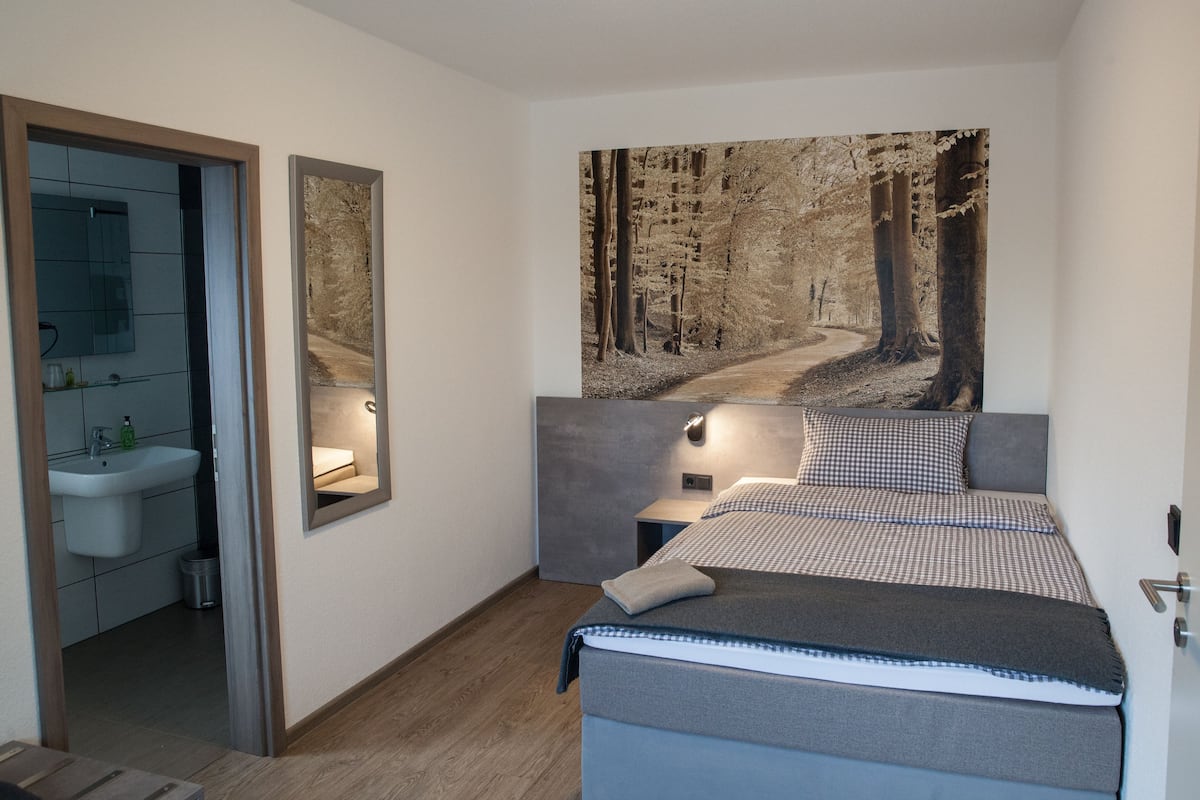 Een slaapkamer met een bed, nachtkastje, spiegel en een groot wanddecor van een bos.