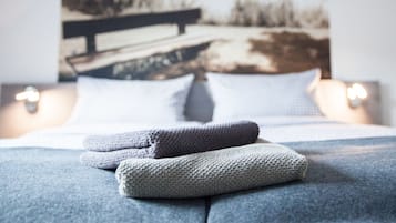 A neatly made bed with a blanket and pillows, a painting of a landscape on the wall behind, and a bedside lamp.