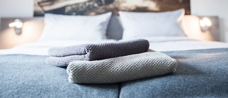Una cama bien tendida con una colcha y almohadas, un cuadro de un paisaje en la pared de atrás y una lámpara de noche.