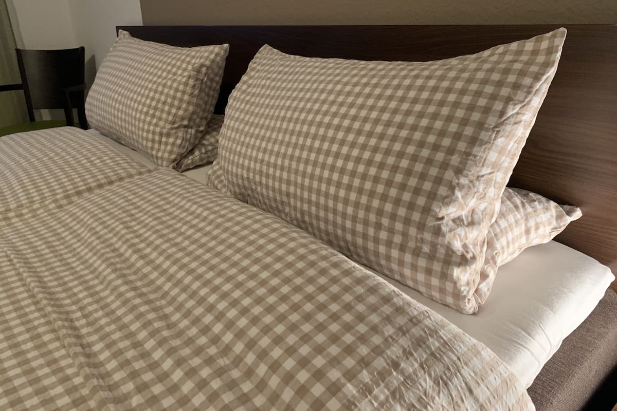 Een opgemaakt bed met geruit beddengoed en kussens, een houten hoofdbord en een effen muur op de achtergrond.