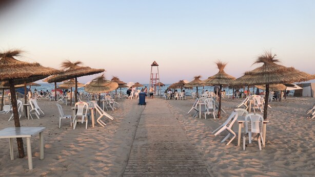 A beach with rows of white plastic chairs and thatched umbrellas, a wooden walkway, and a lifeguard tower in the distance. - Tazarka House (Korba)