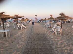 A beach with rows of white plastic chairs and thatched umbrellas, a wooden walkway, and a lifeguard tower in the distance. - Tazarka House (Korba)
