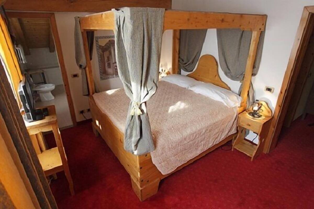 A wooden four-poster bed with a red carpet, a bedside table with a lamp, and a small wooden chair.