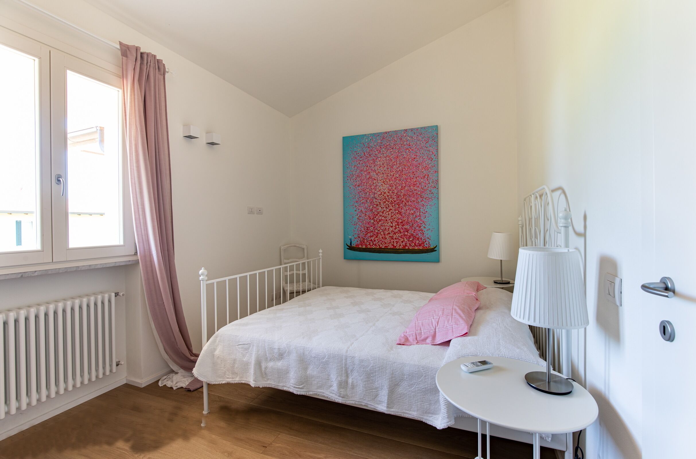 A bedroom with a bed, a bedside table, a radiator, a window with curtains, and a wall-mounted lamp.