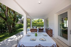 An outdoor patio with a table set for dining, surrounded by wicker furniture and a glass door leading to a garden.
