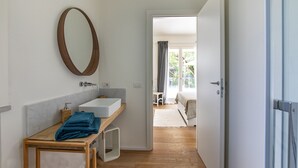 Ein Badezimmer mit einem Holzkosmetikschrank, einem Spiegel und einem weißen Waschbecken. Eine Tür führt zu einem Balkon mit Blick auf Grünflächen.