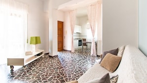 A modern living room with a patterned floor, a sofa with cushions, a small table with a lamp, and a view into the kitchen through a doorway. - Villa Franca (Pietrasanta)