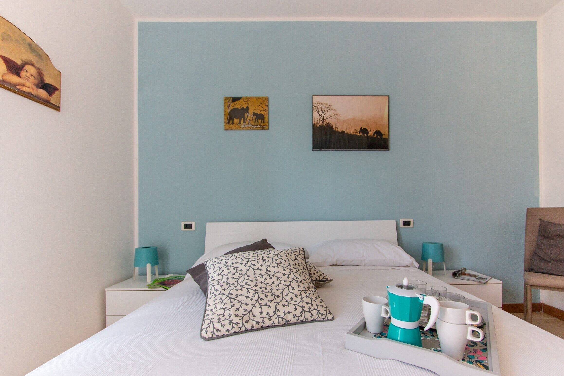 A bedroom with a bed, two pillows, a bedside table with a lamp, a tray with cups and a coffee pot, and two wall art pieces.