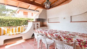 An outdoor patio with a table set for dining, surrounded by white chairs, a stone bench with yellow cushions, and a rustic stone fireplace. - Villa Gloria (Pietrasanta)