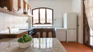 A kitchen with wooden cabinets, a granite countertop, a sink, a stove, a refrigerator, and a window with curtains. - Villa Gloria (Pietrasanta)