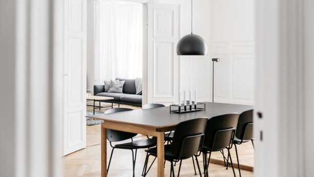 Ein moderner Essbereich mit einem Holztisch, schwarzen Stühlen, weißen Wänden und einem grauen Sofa im Hintergrund.
