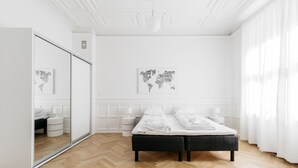 Ein schlicht eingerichtetes Schlafzimmer mit Holzdielen im Fischgrätmuster, einem Bett mit weißer Bettwäsche, einem großen Spiegel und zwei an der Wand hängenden Kunstwerken, die Karten zeigen.