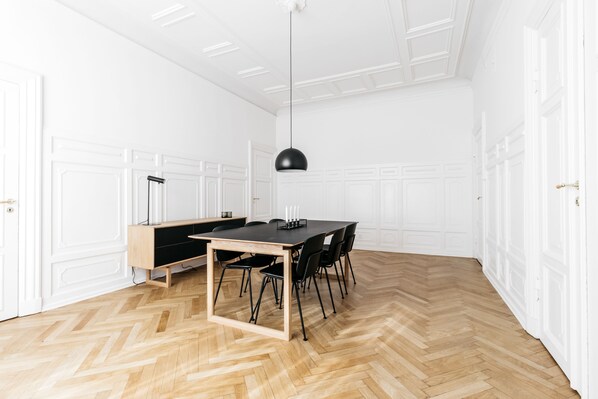 A spacious room with a wooden herringbone floor, white paneled walls, a black pendant light, and a wooden table with chairs.