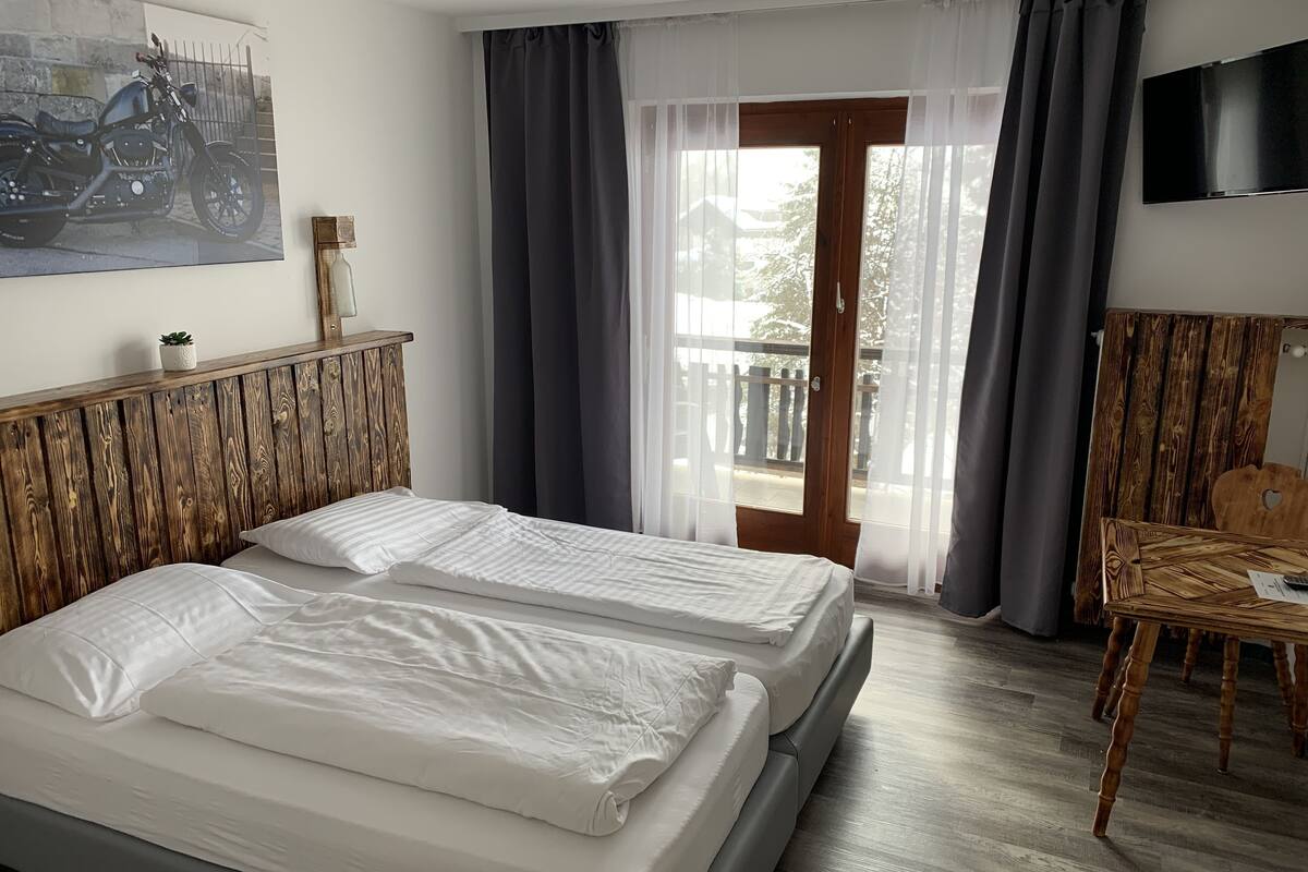 A hotel room with a bed, a wooden headboard, a table, a TV, and a balcony with curtains.
