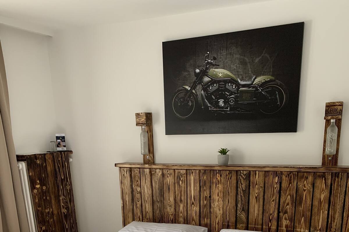 A double bed with white bedding, a wooden headboard, a mounted motorcycle artwork, and a small potted plant on the dresser.