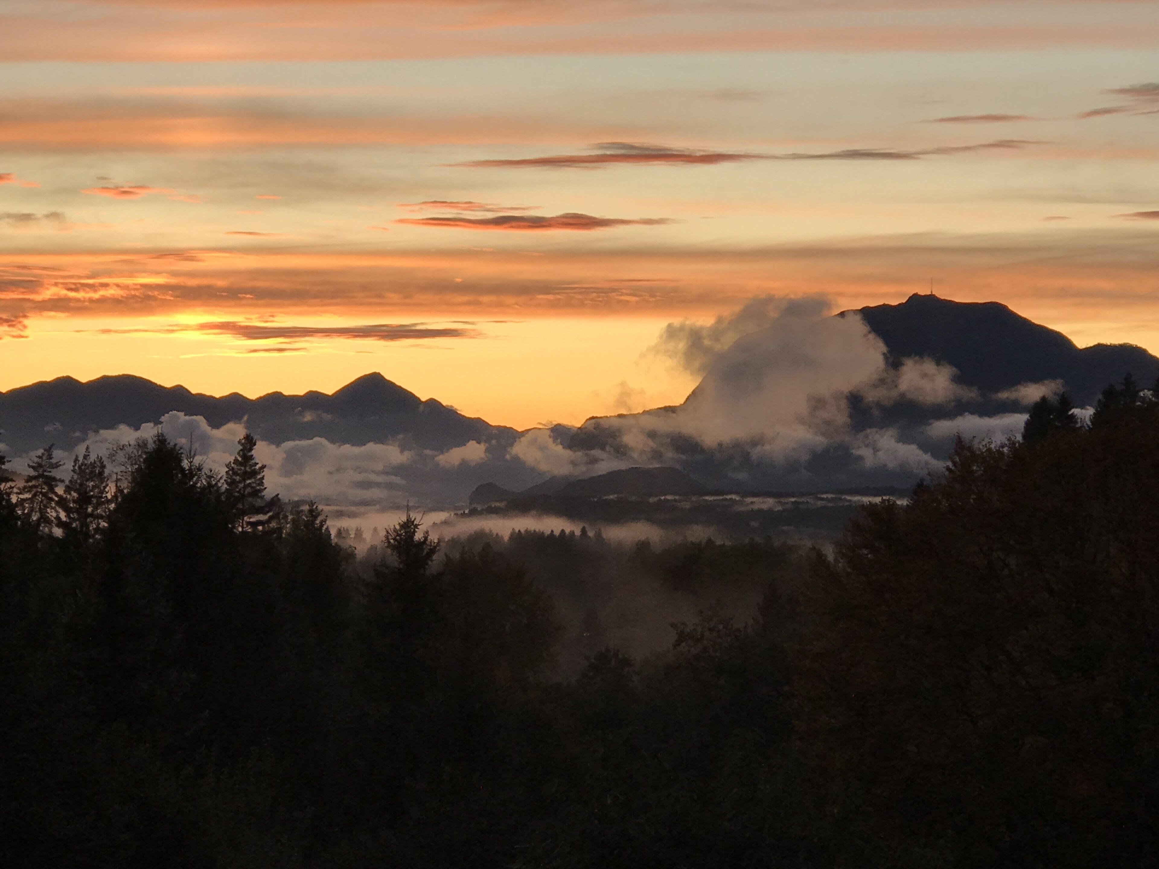 A mountain range at sunset with clouds and trees in the foreground.