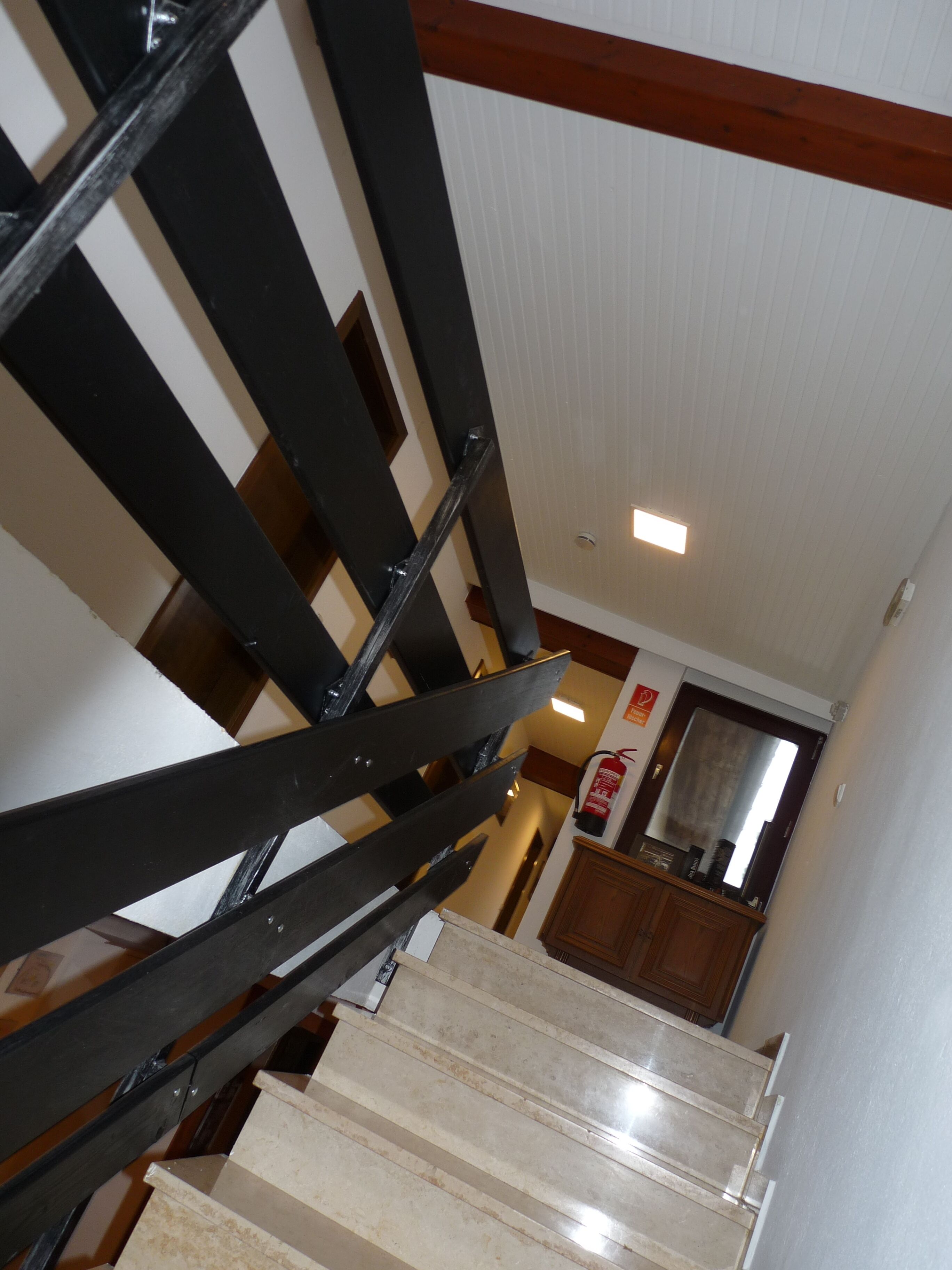 Eine Treppe mit schwarzen Spindeln und beigen Stufen, die zu einem Zwischenpodest mit einem Holzschrank und einem Feuerlöscher führt.