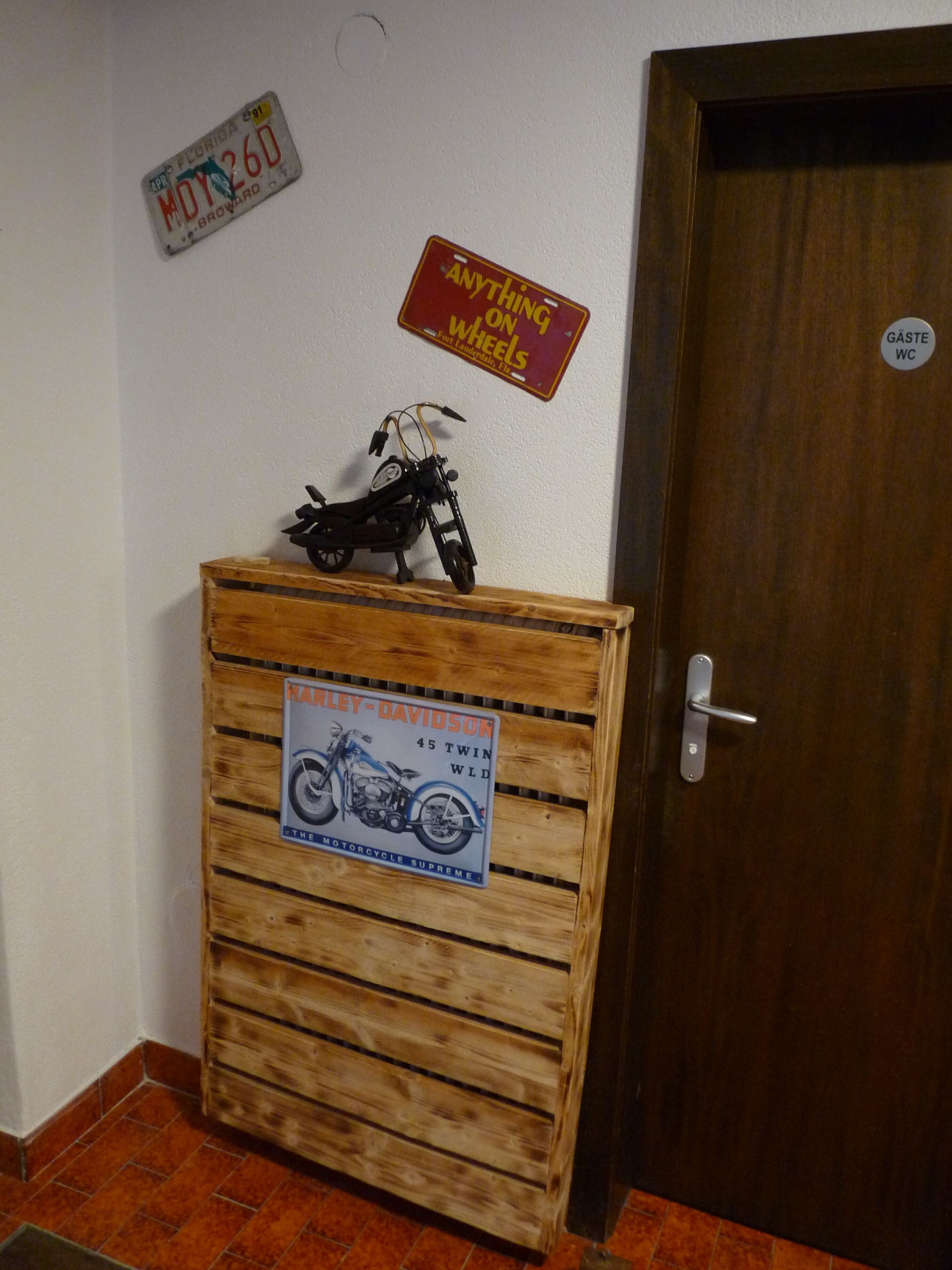 Een houten kast met een motorstickerset, een Florida nummerplaat en een deur met een bordje 'Gasten toilet'.