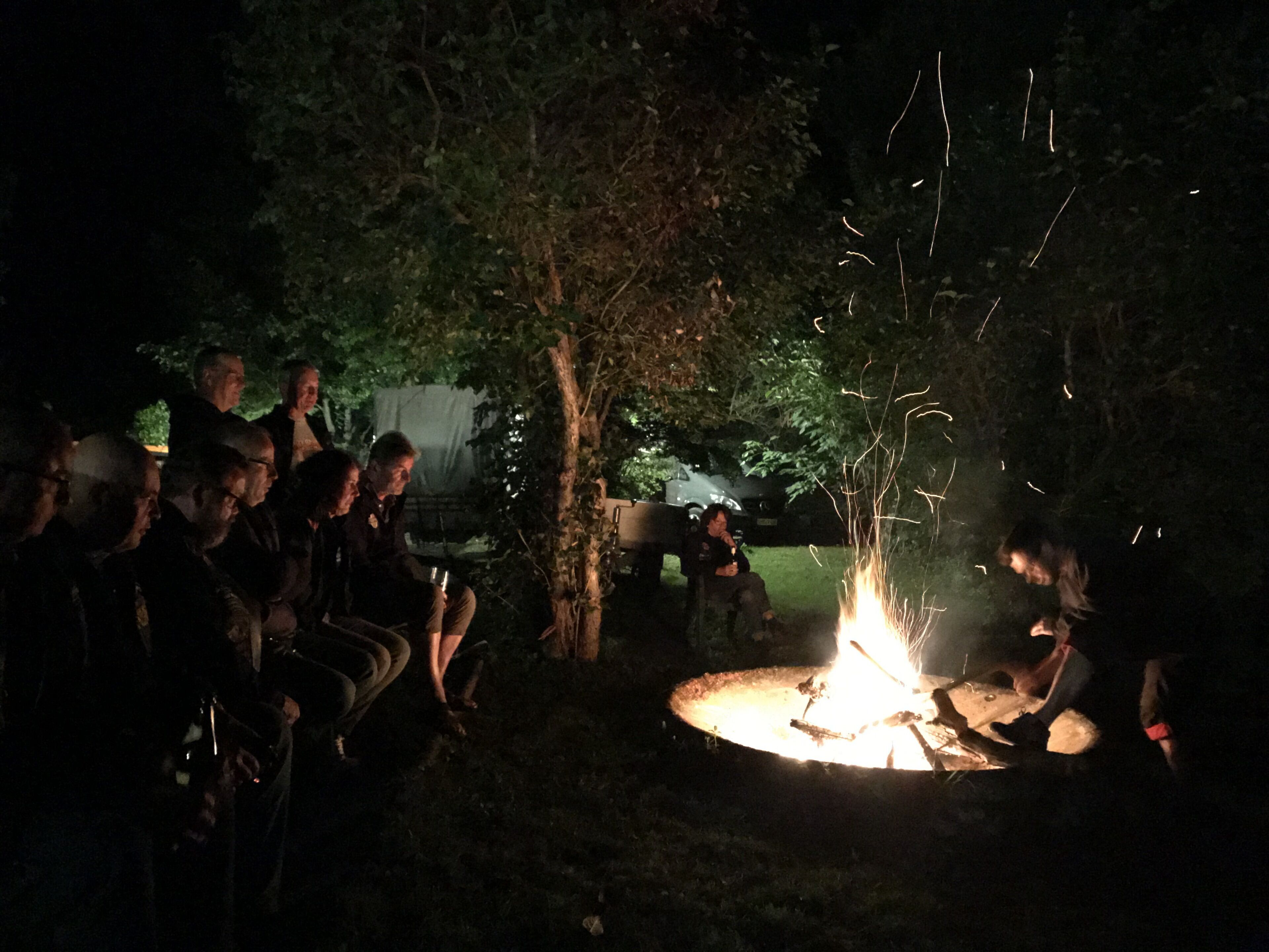 Eine Gruppe von Menschen versammelt sich nachts bei einem Lagerfeuer in einem Waldgebiet.