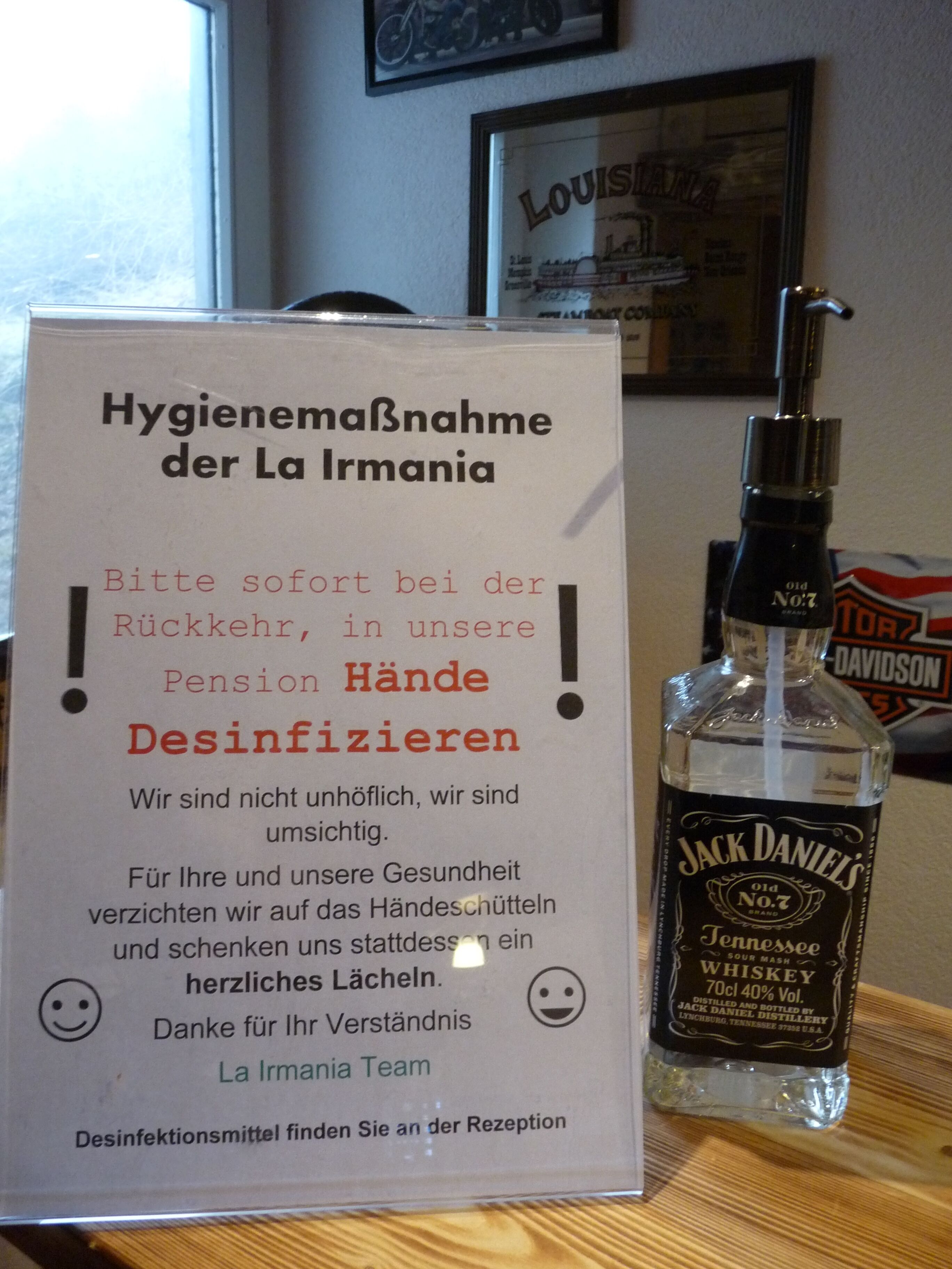 Ein Schild in deutscher Sprache zu Hygienemaßnahmen, eine Flasche Jack Daniel's Whiskey und ein gerahmtes Bild an der Wand.