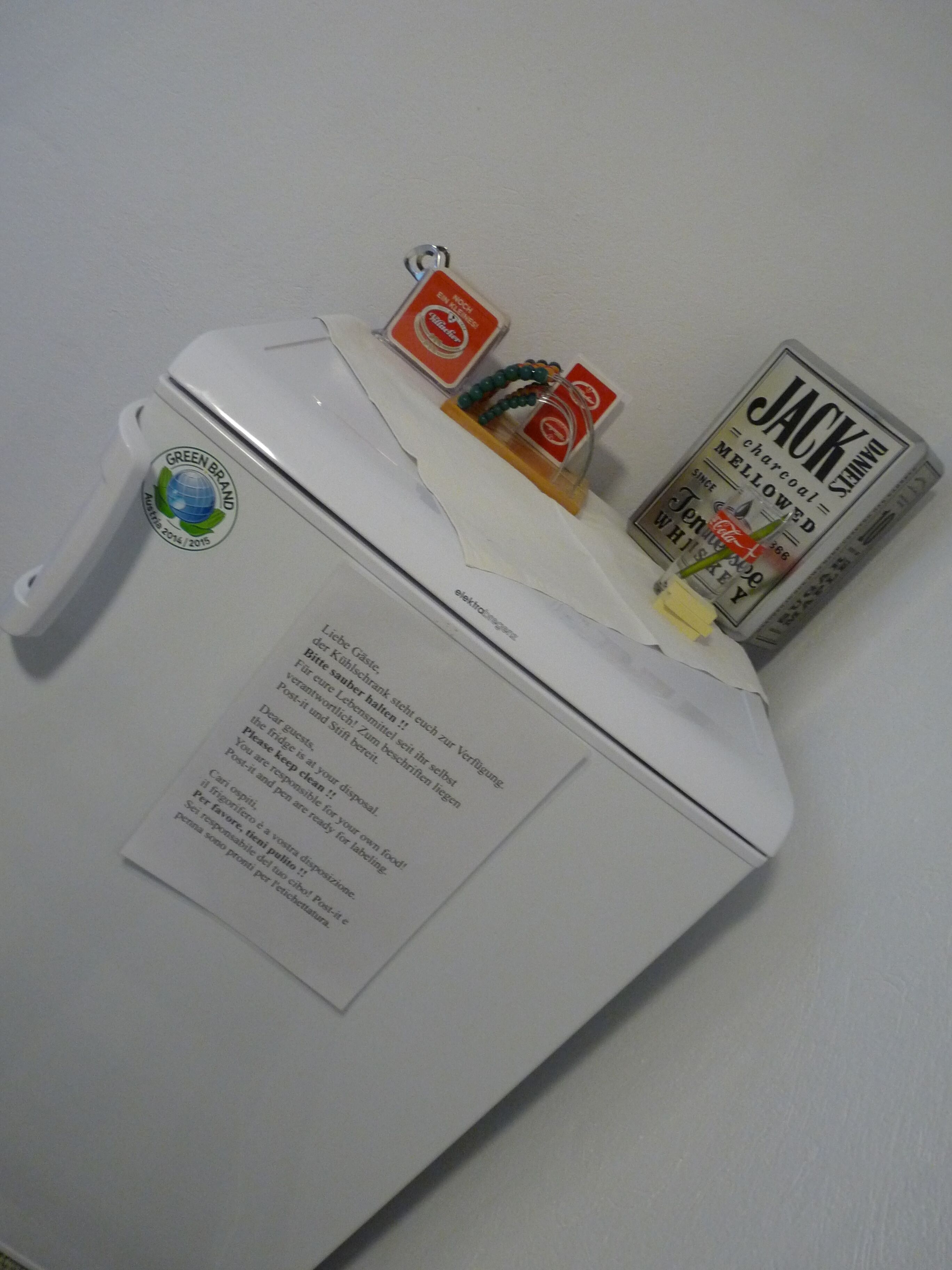A white refrigerator with a green brand sticker, a Jack Daniel's whiskey bottle, and a small beaded necklace.