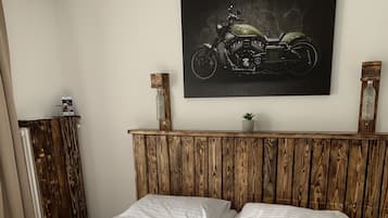 A double bed with white linens, a wooden headboard, a mounted motorcycle artwork, and a small potted plant.