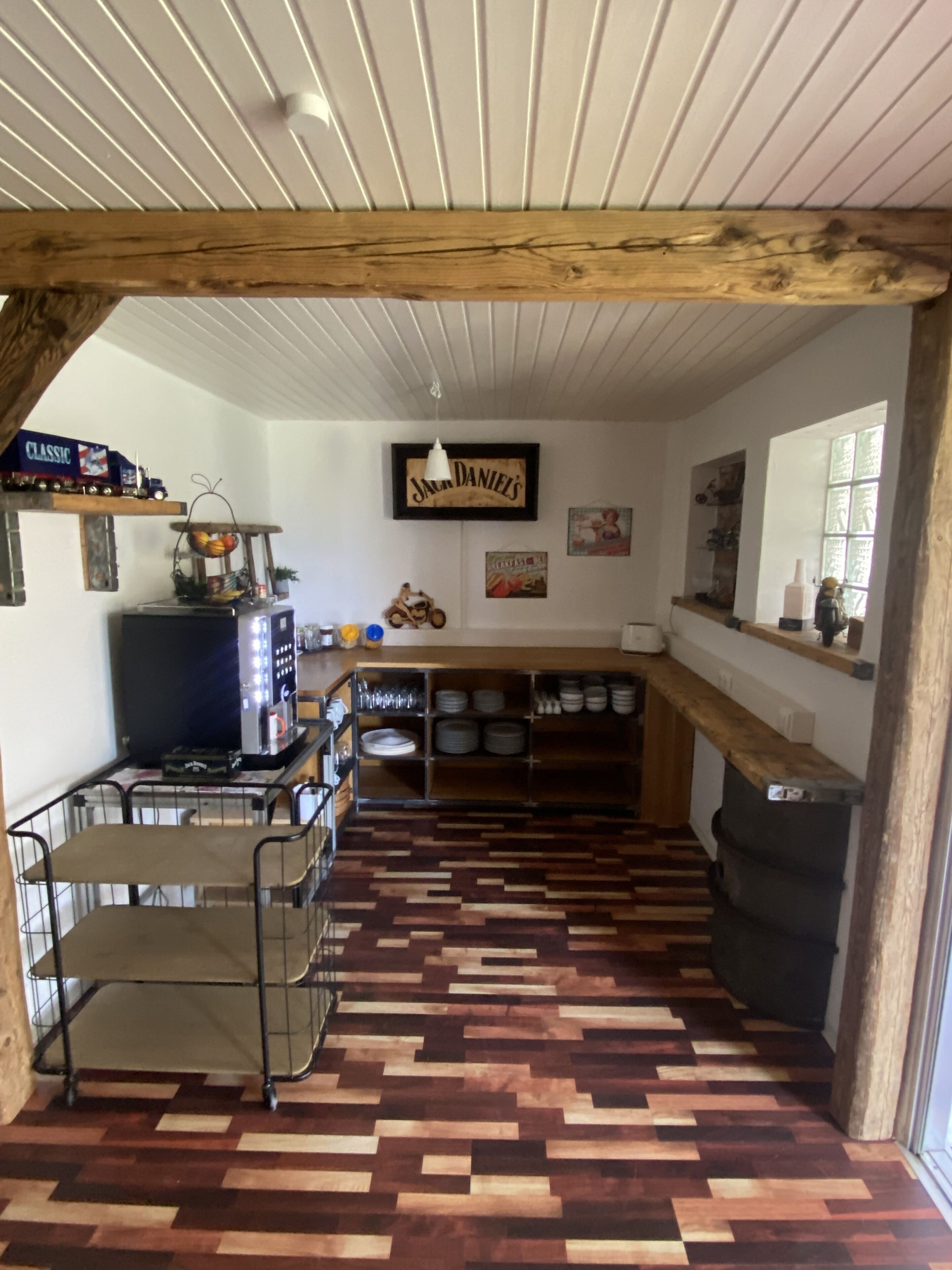 Una cocina con suelo de espiga, vigas de madera, una estufa, repisas con vajilla y una lámpara colgante.