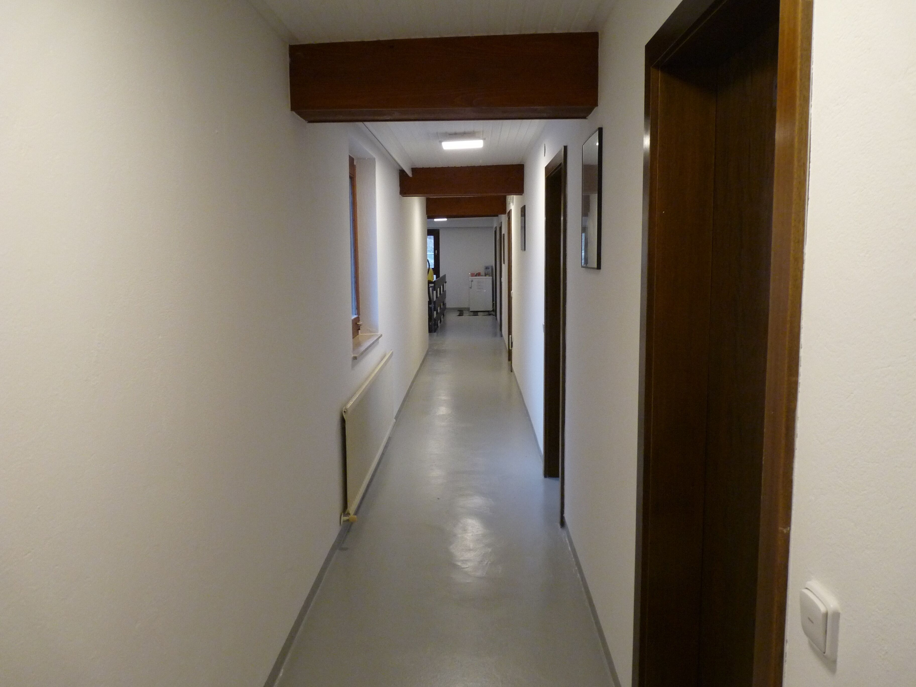 Um corredor longo e estreito, com paredes brancas e chão cinzento, que conduz a um quarto com porta de madeira.