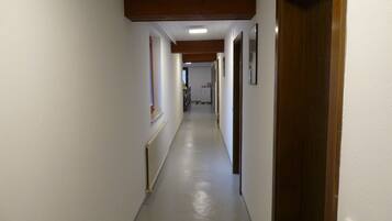 A long, narrow hallway with white walls and a grey floor, leading to a room with a wooden door.