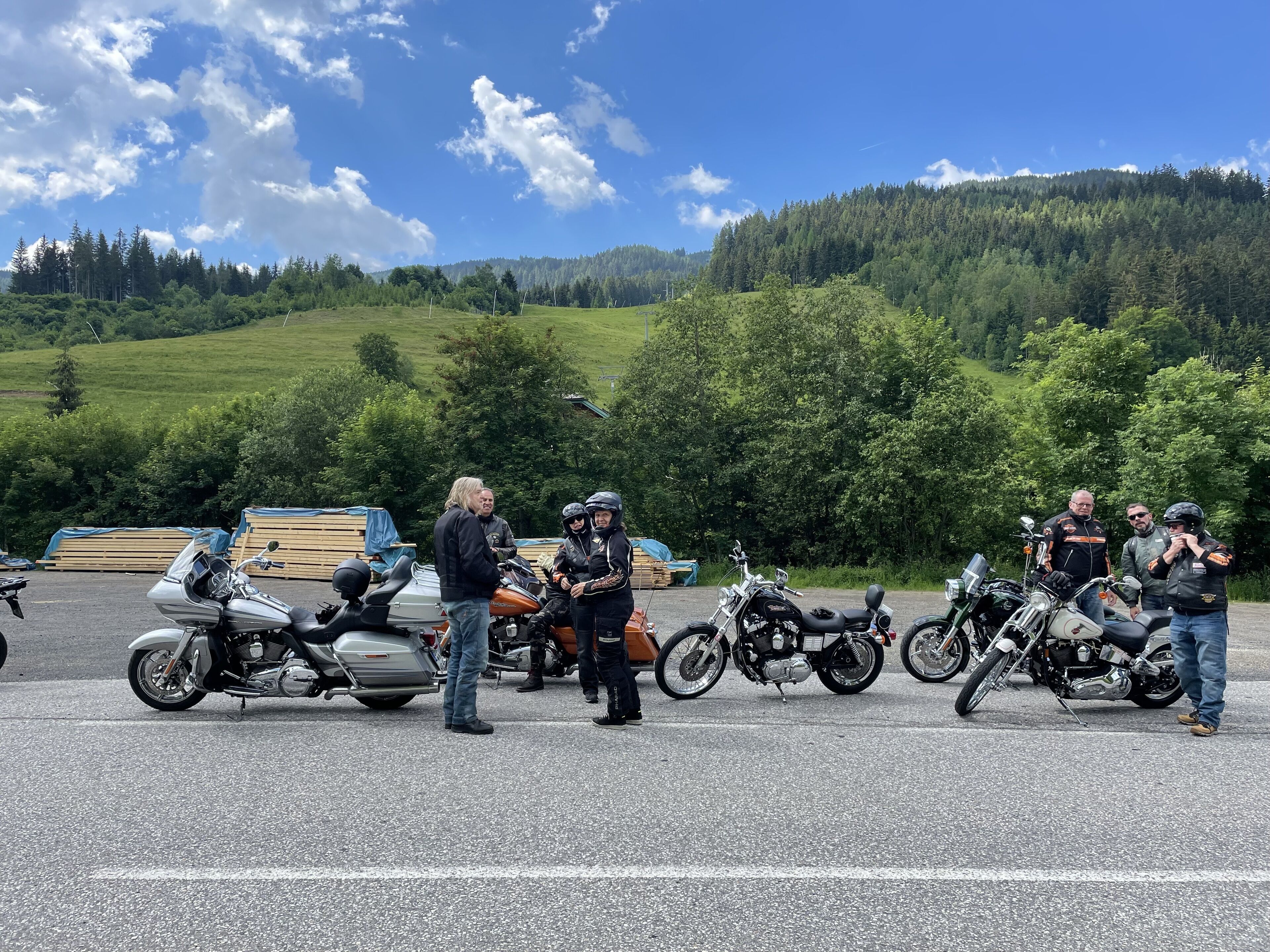 Un grupo de personas cerca de motocicletas en un estacionamiento, con vistas panorámicas a colinas verdes y un cielo despejado de fondo.