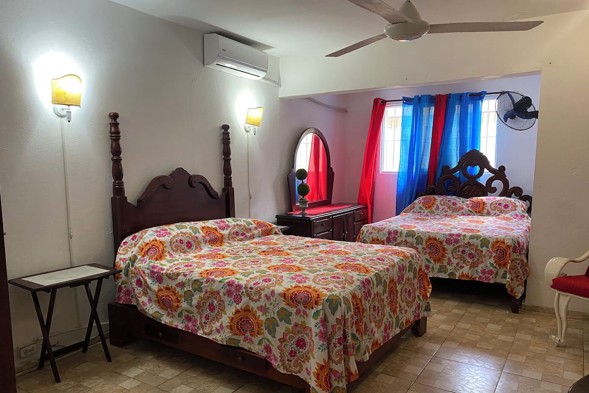 A bedroom with two beds, a ceiling fan, an air conditioner, a nightstand, a mirror, and a window with curtains.