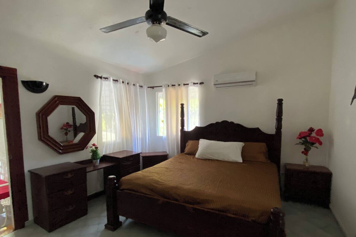 A bedroom with a wooden bed, a ceiling fan, a mirror, a dresser, and a window with curtains.